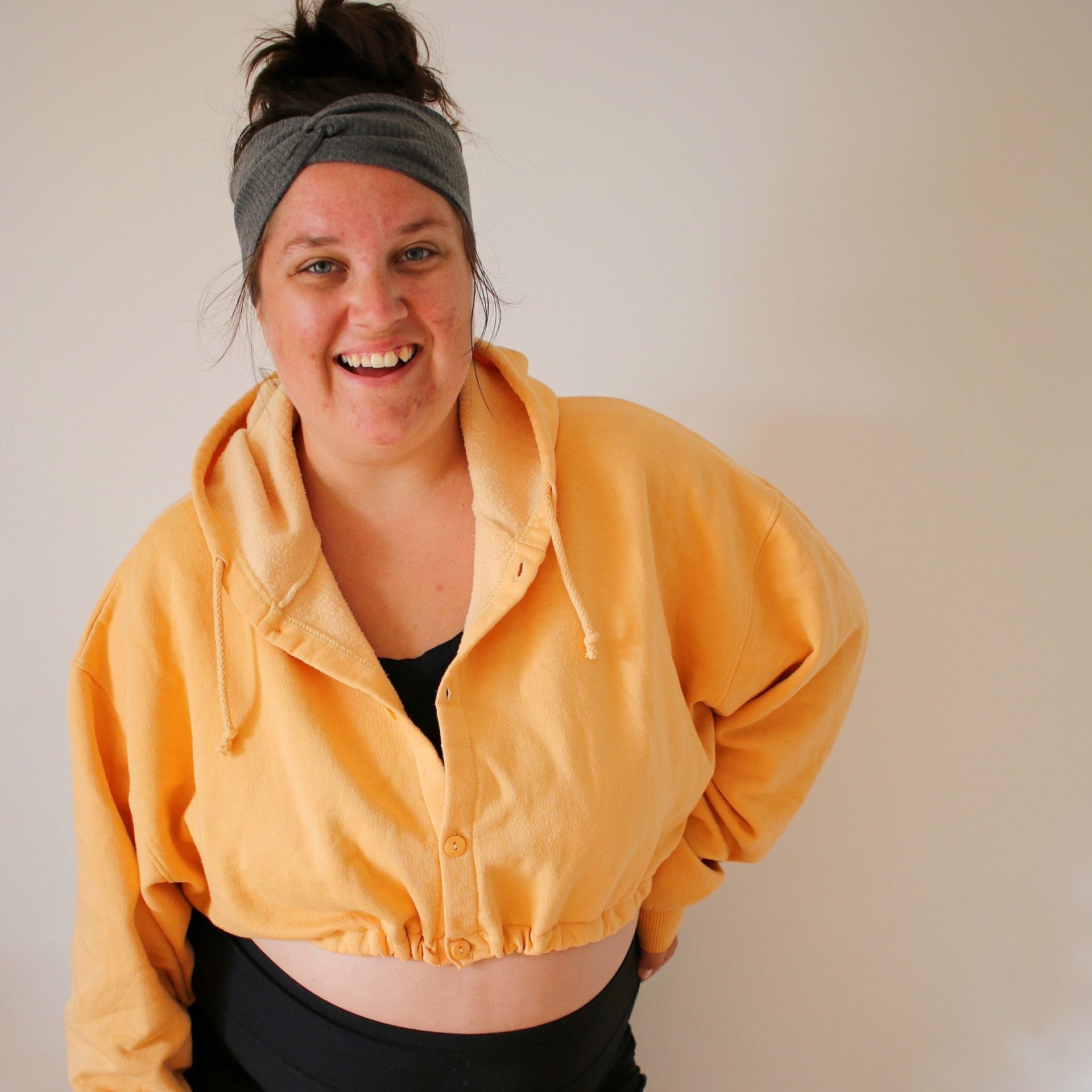 White woman with brown hair up in a bun wearing a grey headband, a golden yellow cropped hoodie and black biker shorts smiles at the camera
